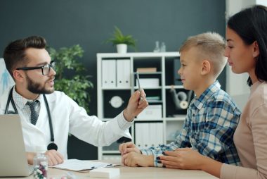 Male pediatrician in white gown is talking to family young mother and son examining child writing in chart during medical appointment in hospital.